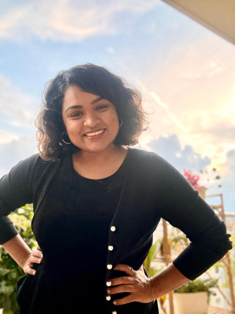 Divya smiling in a black cardigan, standing with her hands on her hips.